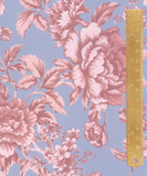 A close-up of a floral fabric pattern featuring large pink peonies and leaves on a dusty blue background, with a gold metal ruler standing vertically on the right side for scale.