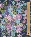 A close-up of a floral fabric pattern with large pink, blue, and purple flowers on a dark navy background, shown with a wooden ruler along the right edge for scale.