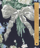 A close-up of a fabric pattern featuring a large white satin ribbon bow tied around a bouquet of white and light blue flowers, set against a dark navy background. A gold metal ruler is positioned vertically on the right side to show scale.