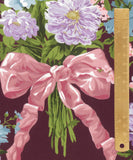A close-up of a floral pattern featuring large light purple flowers, green leaves, and a prominent pink ribbon bow, with a wooden ruler along the right side for scale.