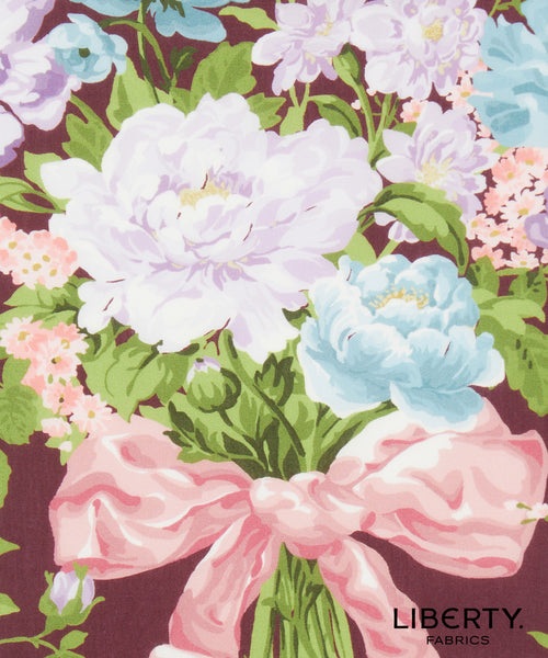 A floral fabric print featuring large white and light blue peonies, pink blossoms, green leaves, and a pink ribbon bow on a dark mauve background. The Liberty Fabrics logo is visible in the bottom right corner.