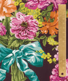 A close-up of a floral fabric pattern with large pink and orange flowers, green leaves, yellow blossoms, and a teal ribbon bow on a dark background, with a wooden ruler along the right edge for scale.