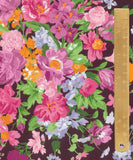 A close-up of a floral fabric pattern with large pink and magenta flowers, green leaves, and small orange and lavender blossoms on a dark background, with a wooden ruler along the right edge for scale.