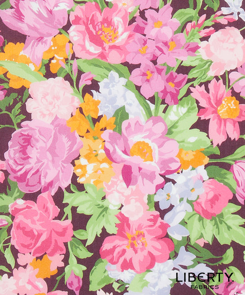 A close-up of a floral fabric print featuring large pink and light pink flowers, green leaves, and small yellow and white blossoms on a dark plum background. The word 'LIBERTY' appears in the lower right corner, with 'FABRICS' below it.