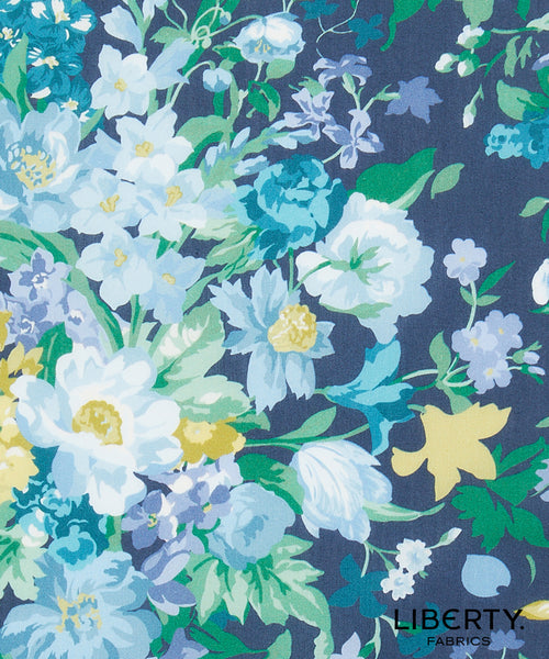 A close-up of a floral fabric print featuring white, light blue, and yellow flowers with green leaves on a dark navy blue background. The Liberty Fabrics logo is visible in the bottom right corner.