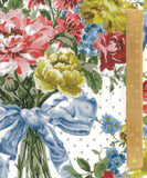 A close-up of a floral fabric print featuring pink, red, yellow, and blue flowers with green leaves, a light blue ribbon, and small gray dots on a white background, with a wooden ruler along the right edge for scale.